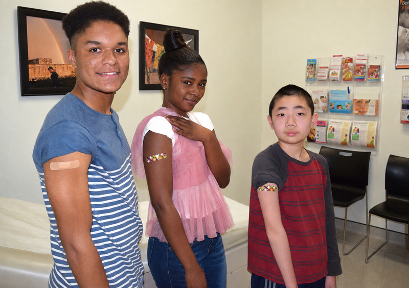 Three children facing the camera showing their arms with a Band-Aid on it from an immunization in a health exam room