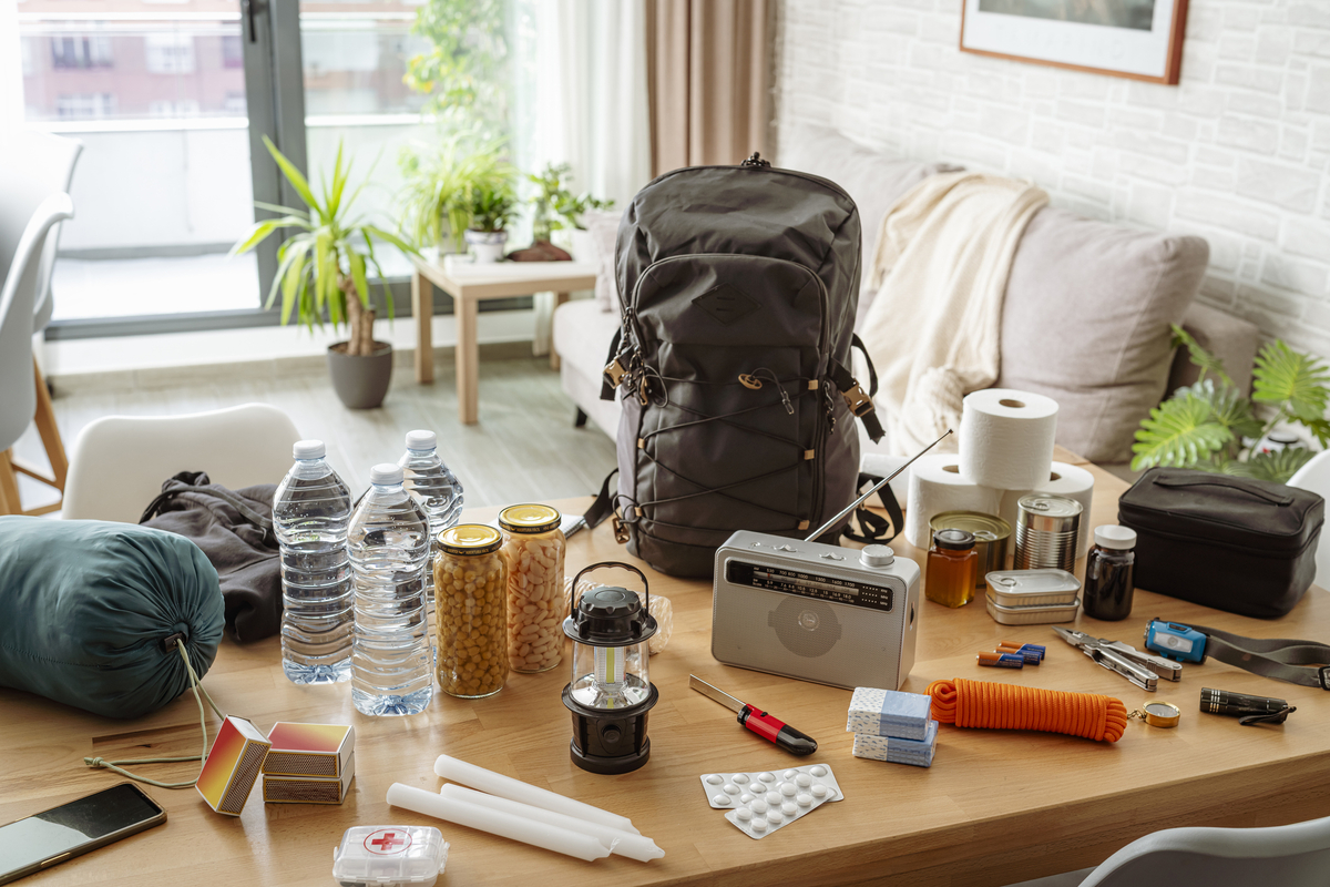 A backpack and emergency supplies are arranged on a table in a bright living room, including bottled water, canned and packaged food, a lantern, radio, first-aid items, batteries, tools, rope, and other preparedness gear.