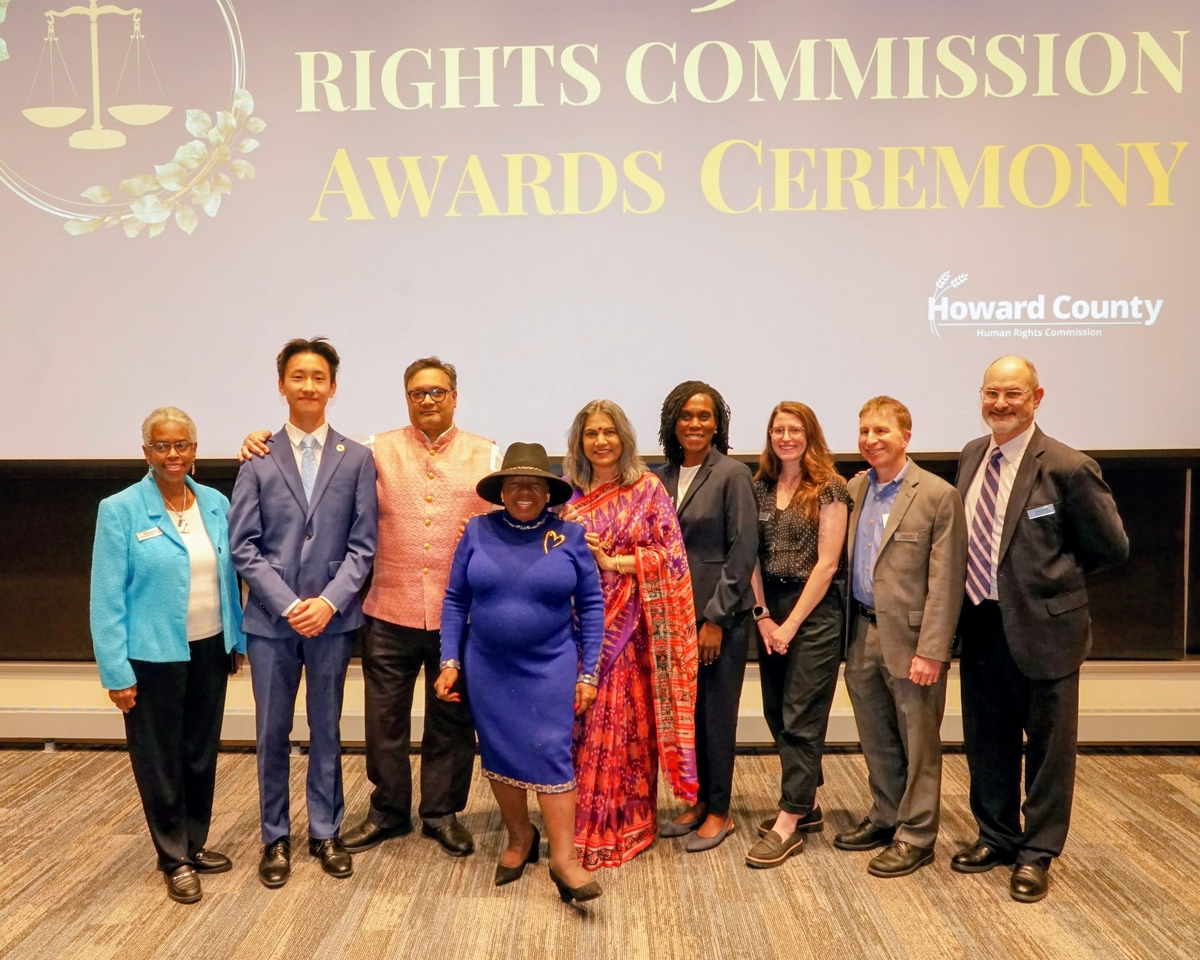 9 people in business formal dress stand in front of a projection reading "2025 Human Rights Commission Awards Ceremony"