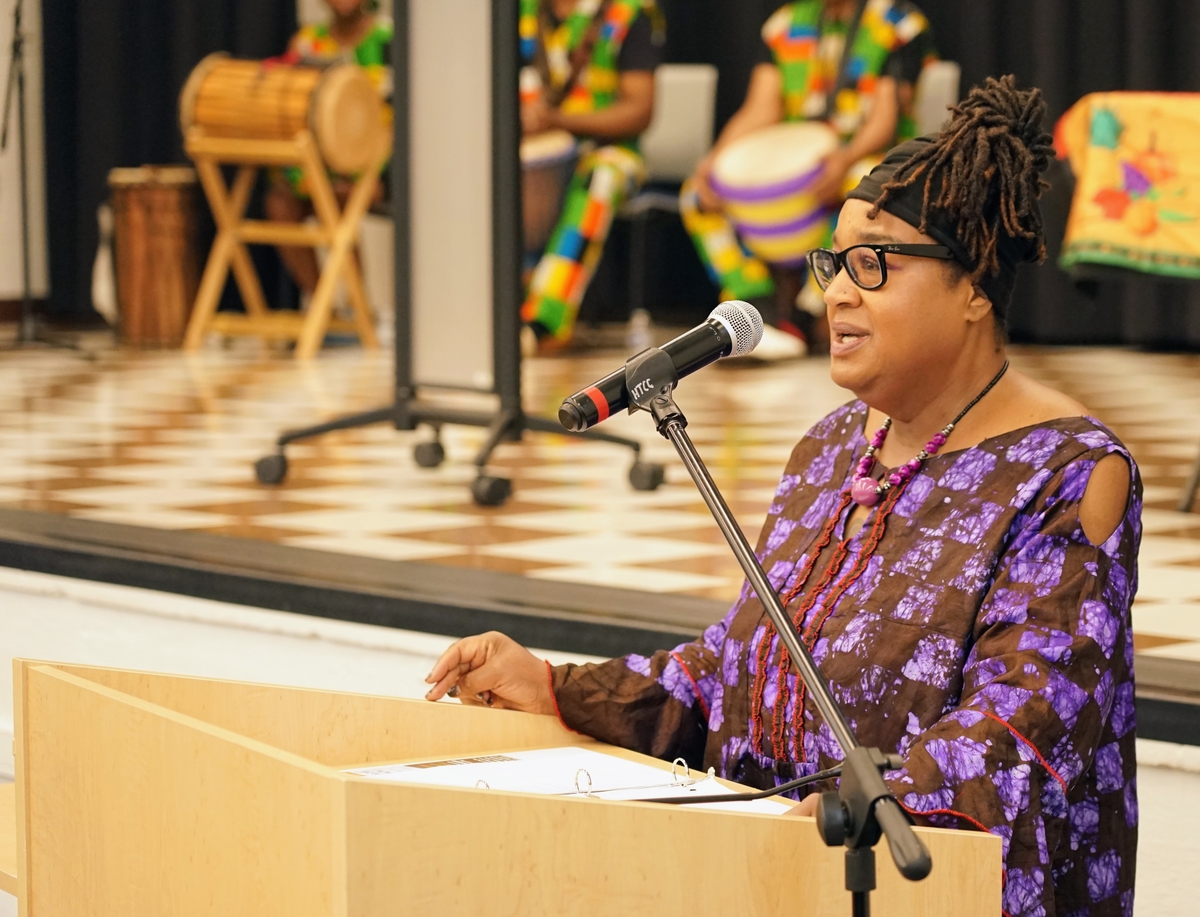 A woman with locs and black glasses speaks into a microphone.