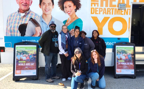 A group of eight people smiling in front of the howard county health department program truck featuring photos of diverse individuals and bold orange text. Two signs on either side of the group read “World AIDS Day,” suggesting the group is participating in or supporting an HIV awareness event.
