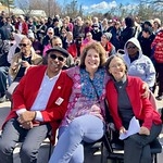 Three older adults in the foreground smiling and a crowd of people in the background.