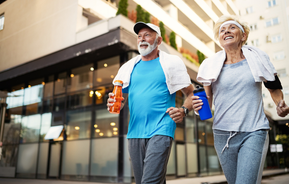 Older adult man and woman walking together in workout clothes holding water bottles and towels around neck.