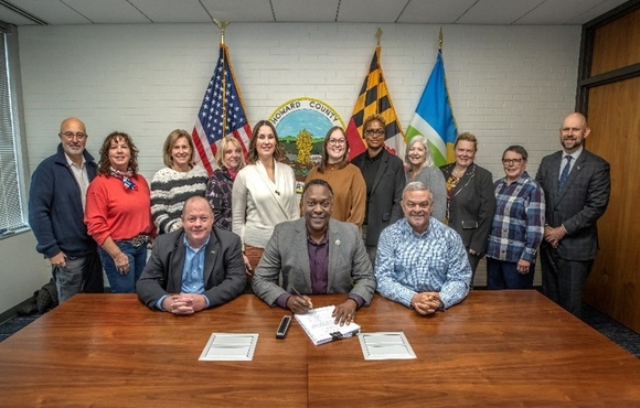 A photo of County Executive Calvin Ball seated at a table with Office of Agriculture Administrator James Zoller and District 5 Councilmember David Yungmann signing into law CB74-2025 ZRA-217 with other staff members lined up behind them.