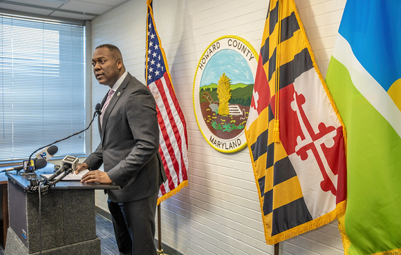 A photo of County Executive Calvin Ball standing at a podium inside his conference room in the George Howard Building, with the County seal and United States, Maryland, and Howard County flags behind him during his briefing to the media about Howard County Government revoking a building permit for a proposed detention center in Elkridge.