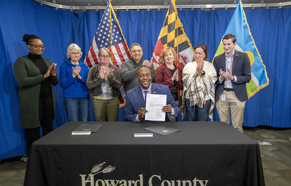A photo of County Executive Calvin Ball seated at a table holding up Council Bill 11-2026 after signing it surrounded by staff and community members.