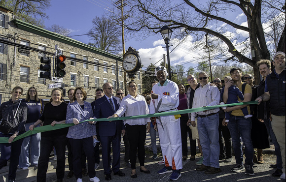 County Executive Calvin Ball holding a giant pair of scissors ready to cut the green ribbon celebrating the unveiling of Old Ellicott City's new clock while posed with fellow elected officials, members of the Ellicott City Partnership, and Old Ellicott City community members.