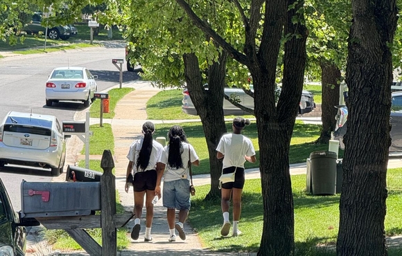 Three youth walk on a sidewalk with a mailbox and several cars visible