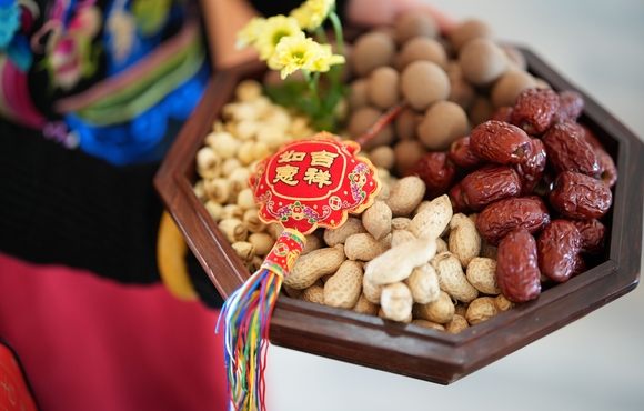 hands carrying a dish of mixed nuts with a red Chinese streamer on top of the one side of nuts (to the viewer's left)