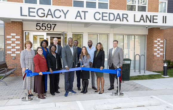 County Executive Calvin Ball posed with others standing outside the front entrance to Legacy at Cedar Lane II housing complex and in front of a blue ribbon ready to celebratory cut the ribbon to mark the projects completion and opening.