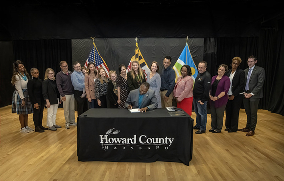 County Executive Calvin Ball seated at a table signing Council Resolution 159-2026 into law as individuals lined up behind him look on. The American, Maryland, and Howard County flags stand in the background.