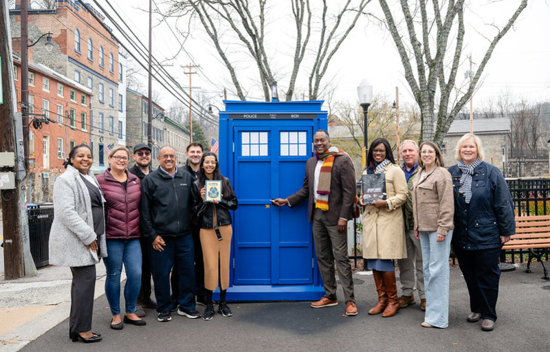 A photo of County Executive Calvin Ball and County staff and local Old Ellicott City community members standing in front of the County's new mobile Little Free Library the #HoCoTARDIS.
