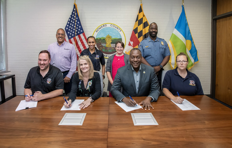 County Executive Calvin Ball seated at a table with the County's Chief Administrative Officer Brandee Ganz and members of Howard County Government and Howard County Public Dispatcher’s Association, with other officials standing behind, signing a memorandum of agreement between the County and the group.