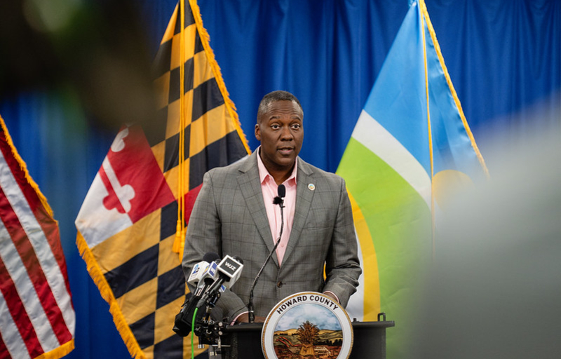 County Executive Calvin Ball speaking at a podium addressing a crowd at his press conference with the American, Maryland, and Howard County flags behind him.