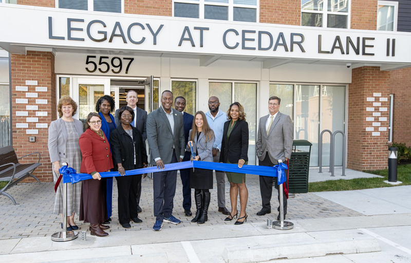 County Executive Calvin Ball posed with others standing outside the front entrance to Legacy at Cedar Lane II housing complex and in front of a blue ribbon ready to celebratory cut the ribbon to mark the projects completion and opening.