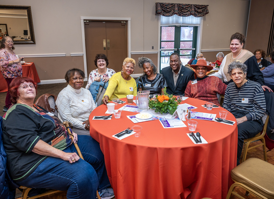 Group of older adults posing for a photo around a round table with orange tablecloth. The County Executive and OAI Administrator are also in the photo.