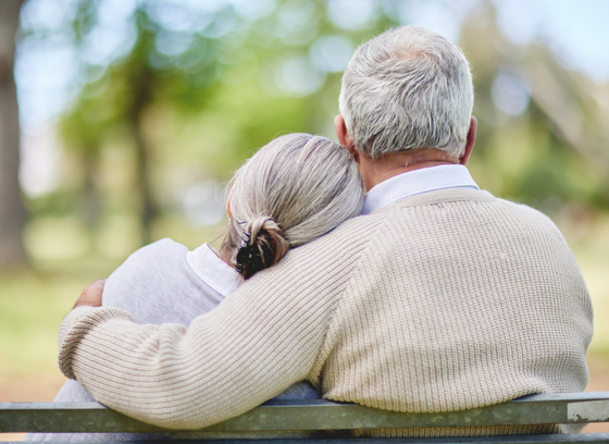 Older couple with grey hair sitting on bench with arms around each other.