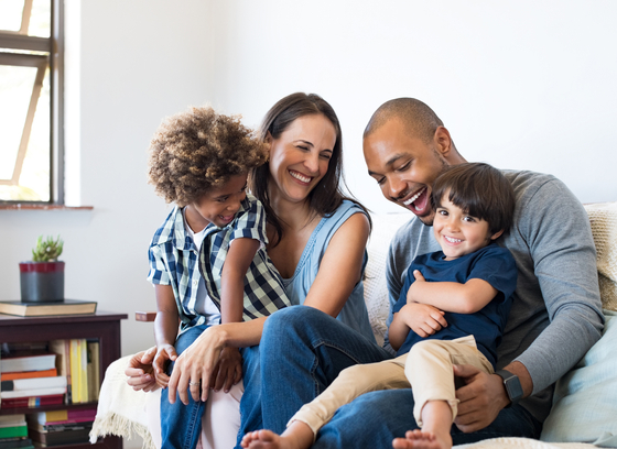 A smiling family of four sits together on a light-colored couch in a bright living room. Two adults and two young children are laughing and cuddling close to one another. Sunlight streams in through a large window on the left, and a small table with books and a potted plant is visible in the background.