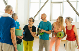 Group of 6 older adults in exercise outfits and some holding yoga mats. There are 2 men and 4 women. They look happy and chatting with each other.