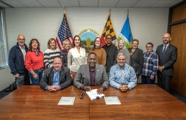 A photo of County Executive Calvin Ball seated at a table with Office of Agriculture Administrator James Zoller and District 5 Councilmember David Yungmann signing into law CB74-2025 ZRA-217 with other staff members lined up behind them.
