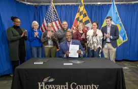 A photo of County Executive Calvin Ball seated at a table holding up Council Bill 11-2026 after signing it surrounded by staff and community members.