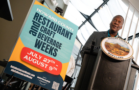 County Executive Calvin Ball speaking at a podium inside River House on Main with a poster board set up next to him with information about Howard County Restaurant & Craft Beverage Weeks for summer 2026, running from July 27th to August 9th.