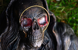 A close-up of a dark, weathered skull figure wearing a black hood, featuring red-painted eye sockets and a thin gold ring encircling its face against a green grass background.