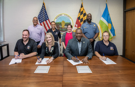 County Executive Calvin Ball seated at a table with the County's Chief Administrative Officer Brandee Ganz and members of Howard County Government and Howard County Public Dispatcher’s Association, with other officials standing behind, signing a memorandum of agreement between the County and the group.