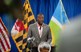 County Executive Calvin Ball speaking at a podium addressing a crowd at his press conference with the American, Maryland, and Howard County flags behind him.