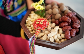 hands carrying a dish of mixed nuts with a red Chinese streamer on top of the one side of nuts (to the viewer's left)