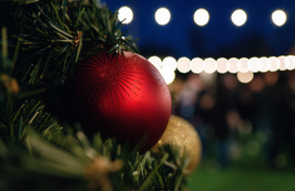 green christmas tree branches in the foreground adorned with a gold ball and a red ball ornament and white string lights glowing the background