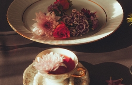 a small white teacup filled with red and white flowers on a white saucer sitting surrounded by a string of pearls near a bowl filled with white, red, and brown flowers all on a brown wooden table adorned with flower petals
