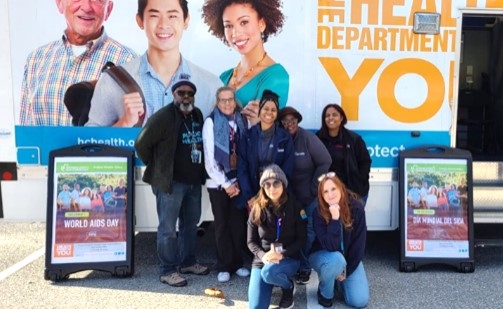 A group of eight people pose together outdoors in front of a large health department mobile unit. The vehicle features photos of smiling individuals and bold text related to health services. Two signs on either side of the group display messages about World AIDS Day. The group is casually dressed, smiling, and standing and crouching in a friendly formation on a sunny day.