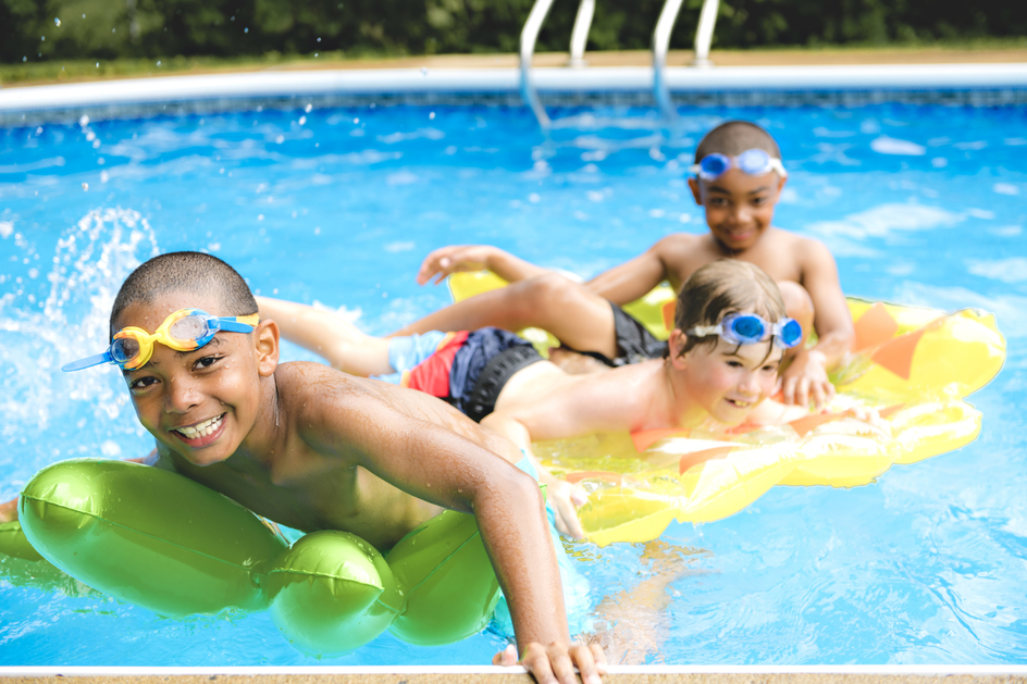 This image shows three children playing in an outdoor swimming pool, wearing goggles and using colorful inflatable floats in a sunny, relaxed setting.