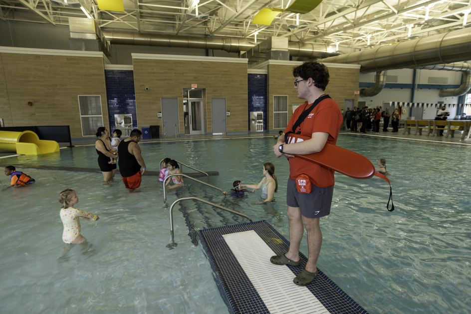 An indoor swimming pool where a lifeguard in a red shirt stands on a platform holding a rescue float, watching over several children and adults playing and wading in the water, with a small slide and seating area visible in the background.