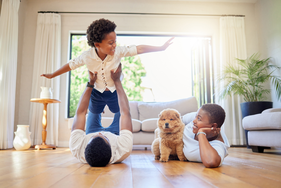 A family of four and a dog laying on the floor together while the man is holding the child in the air in a playful manner