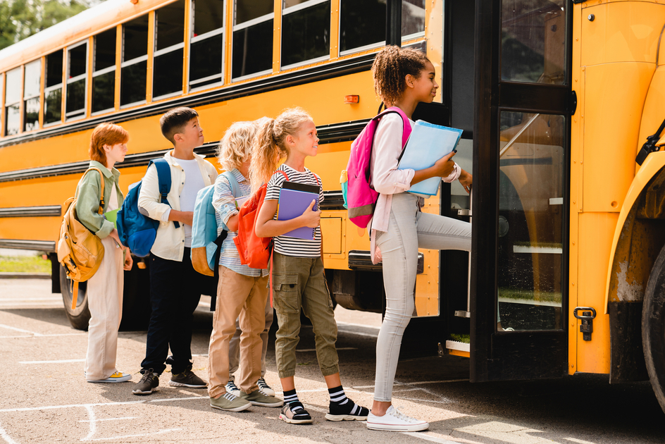 A line of schoolchildren wearing backpacks waits to board a yellow school bus, with the first child stepping onto the bus while holding notebooks.