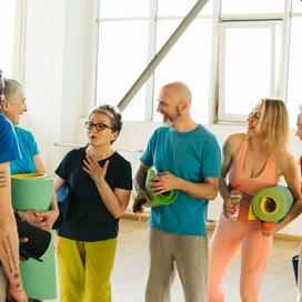 Group of 6 older adults in exercise outfits and some holding yoga mats. There are 2 men and 4 women. They look happy and chatting with each other.