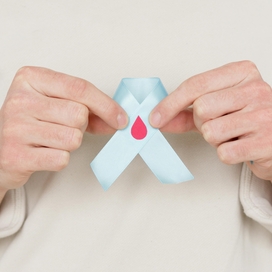 Caucasian person holding up light blue ribbon with blood droplet in their hands. No face shown.