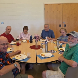Group of eight older adults sitting around a round table eating lunch together and smiling.