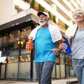 Older adult man and woman walking together in workout clothes holding water bottles and towels around neck.
