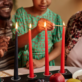 Two adults watch as a child lights a red candle on a Kwanzaa kinara