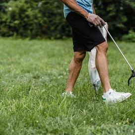 Man picking up trash on grass with a grabber; only abdomen to feet showing.