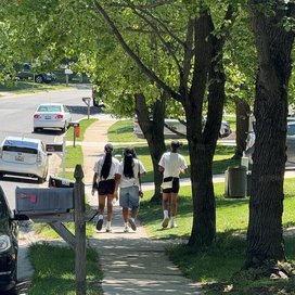 Three youth walk on a sidewalk with a mailbox and several cars visible
