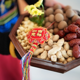 hands carrying a dish of mixed nuts with a red Chinese streamer on top of the one side of nuts (to the viewer's left)