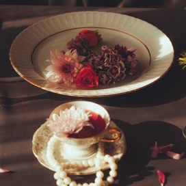 a small white teacup filled with red and white flowers on a white saucer sitting surrounded by a string of pearls near a bowl filled with white, red, and brown flowers all on a brown wooden table adorned with flower petals